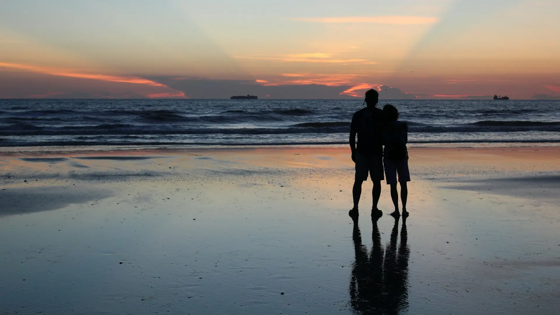 Two people standing close together on wet sand at sunrise, looking out to sea